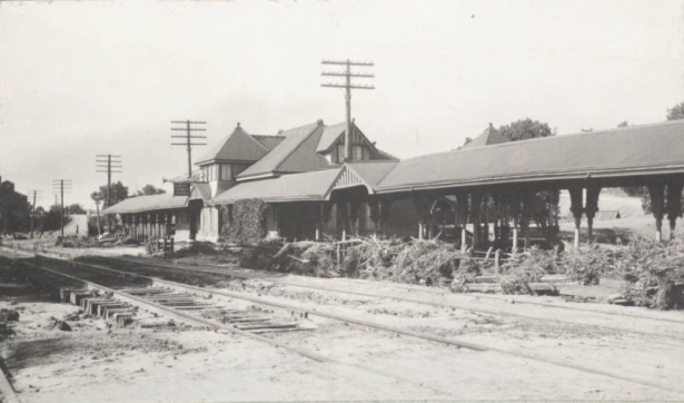View of the 8th (Eighth) Street Depot beside Denver and Rio Grande Western Railroad tracks in Pueblo (Pueblo County), Colorado. Shows mud covered tracks and debris from flooded Fountain Creek washed up beside the covered passenger platform. The frame depot building has a hipped roof, half-timbered dormer windows and a sign that reads: "Eighth Street Pueblo."