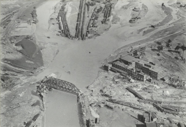 Aerial view of the flooded Arkansas River in Pueblo (Pueblo County), Colorado. Shows freight cars at the washed out Missouri Pacific Railway Company yard, railroad bridges over the river and the Nuckolls Packing Company building beside standing water.