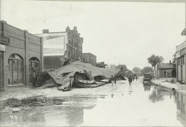 People walk on 2nd (Second) Street in front of damaged commercial buildings and piles of debris from the Arkansas River flood in Pueblo (Pueblo County), Colorado. Men pose on wreckage from a collapsed roof beside a damaged brick building with a decorative parapet. Shows cars, water and mud in the street.
