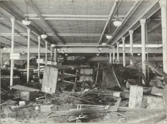 Interior view of White and Davis clothing store at 301 N. Main Street after the Arkansas River flood in Pueblo (Pueblo County), Colorado. Shows wrecked furniture, crates, mud and debris in the aisle. Signs for "Boys Dept." and "Collars" are posted on pipe columns with Corinthian capitals. The store has a pressed metal ceiling and overhead lamps with glass globes.