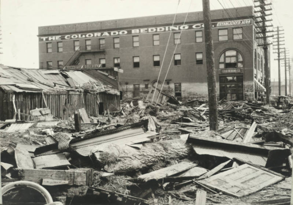 Debris from the Arkansas River flood at 2nd (Second) Street and Grand Avenue in Pueblo (Pueblo County), Colorado. Shows mud, timber, portions of ceiling molding, and a door near the wrecked, frame "Mammoth Corral" building. The three story brick "Colorado Bedding Co." building shows paint chipped off at the high water mark above first story windows.