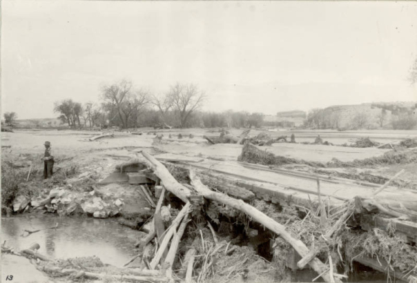 A man in overalls and a fedora stands beside a debris covered Atchison, Topeka and Santa Fe Railroad bridge and a mud covered Denver and Rio Grande Western bridge in probably Pueblo County, Colorado. Tree branches and mud are washed up against the bridge and over the sets of railroad tracks after a flood. Shows an eroded river bed of probably the Arkansas River.