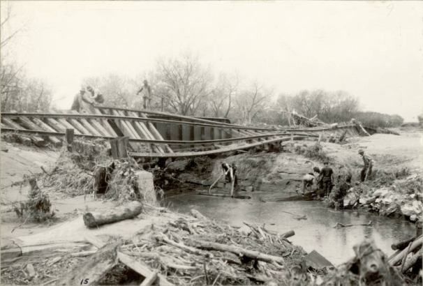 Men work to clear debris from a flood under washed out Denver and Rio Grande Western Railroad bridge number "126-B" in probably Pueblo County, Colorado. Shows tree branches and mud in an eroded creek bed, and unsupported tracks over the creek.