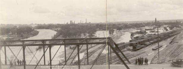 Panoramic view of flood damage from the Arkansas River flood in Pueblo (Pueblo County), Colorado. Shows washed out Denver and Rio Grande Western Railroad tracks and the rail yard near the Union Depot, mud and debris on the ground and smokestacks in the distance. Men stand near a car on the West 4th (Fourth) Street bridge.