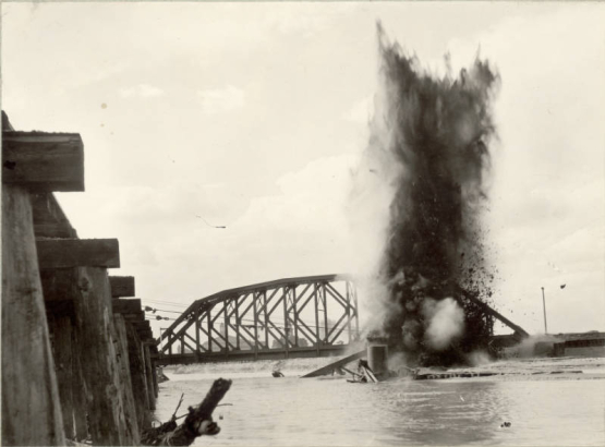 View of a dynamite explosion to dislodge a Denver and Rio Grande Western Railroad bridge from flood debris in the Arkansas River in Pueblo (Pueblo County), Colorado. Shows mud and sand in the air, part of a D. & R.G. bridge submerged in the river and an Atchison, Topeka and Santa Fe Railroad bridge nearby.