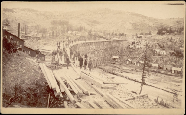 Construction workers in sack suits and overcoats, hold shovels and pose on a Helena and Northern Railroad (a Northern Pacific short line) trestle under construction in Marysville (Lewis and Clark County), Montana. The high, wooden trestle is over a gulch near houses and mine buildings. Piles of lumber are beside the trestle.