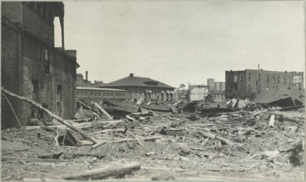 Piles of debris from the Arkansas River flood are in the streets around the stone Atchison Topeka and Santa Fe Railroad depot in Pueblo (Pueblo County), Colorado. Lumber, metal, mud and silt cover the streets near brick commercial buildings.