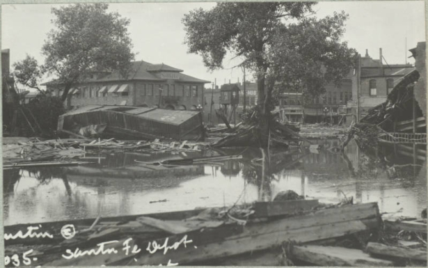 View of overturned freight cars and wreckage from the Arkansas River flood on Union Avenue in front of the stone Atchison, Topeka and Santa Fe Railroad depot in Pueblo (Pueblo County), Colorado. Shows standing water around trees and piles of debris.