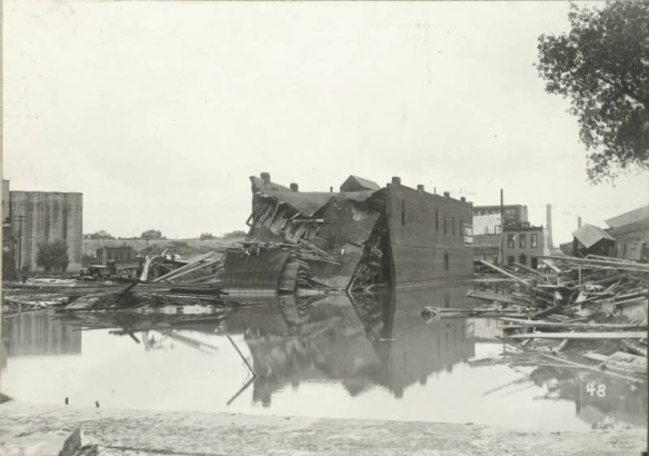 View of standing water piles of lumber and debris on Main Street and Hobson Street after the Arkansas River flood in Pueblo (Pueblo County), Colorado. Shows the collapsed wall of the Ford Exchange building.