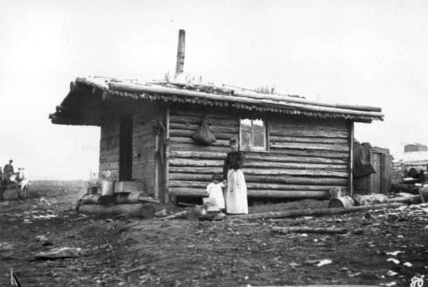 A woman holds a child's hand at the side of a log cabin, Goldfield, Teller County, Colorado. The front gable house is constructed of logs and hewn lumber and has one glass pane window, a mud and sod roof with a stove pipe. Several wooden boxes and crates are piled in front of the cabin and a full sack is hung on the side. A man in a horse-drawn wagon shows in distant far left.