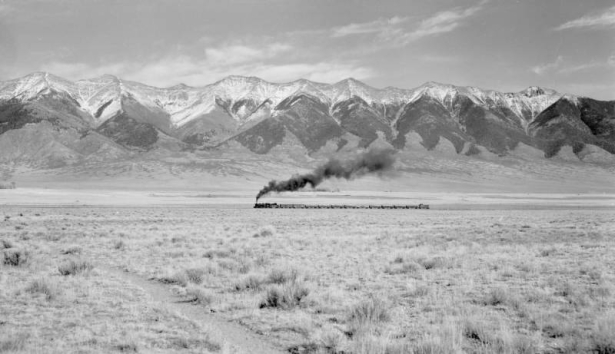 A Denver and Rio Grande Western narrow gauge train pulled by locomotive 480 heads northwest and vents smoke near Villa Grove (Saguache County), Colorado. Clouds and snow top the Sangre De Cristo range.