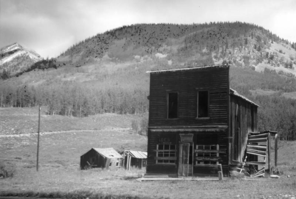 View of a dilapidated two-story wood frame false front with a corrugated metal gable roof, dentiled cornice, two upper-story windows, remains of an outside side stairway and a single door entry flanked by windows in Gothic, Gunnison County, Colorado. A faded sign reads: Lee's Tavern; two small gabled sheds show in background. Gothic was a mining boom town from 1879 to 1884; bought, in 1928, by the Rocky Mountain Biological Laboratory for plant and animal research.
