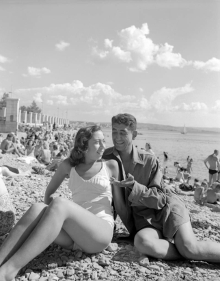 A Tenth Mountain Division soldier enjoys the beach at Trieste with a woman. Both wear bathing costumes and pose for the camera. Behind them the beach is filled with other sun worshipers.