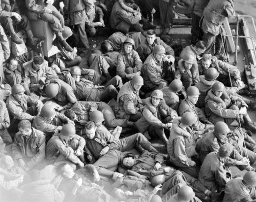 Tenth Mountain Division troops fill the entire frame, except for a small triangle of water in the upper right corner; troops are returning by ship to the U.S.A.  All are in uniform, most wear helmets and some wear life vests. Some are reading, some resting, others look out to sea.