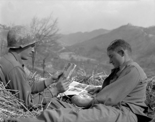 Taken during the Italian campaign, the photograph shows two Tenth  Mountain Division soldiers relaxing in a haystack by reading magazines. In  the background are mountains and leafless trees.