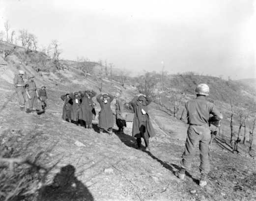 Five Tenth Mountain Division soldiers guard five tagged German prisoners on an Italian hill. Trees in the background are leafless. Photographer's shadow is visible in lower left corner.