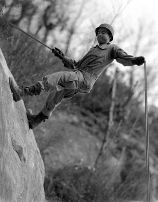 Shows a Tenth Mountain Division soldier wearing a helmet rappelling on a rock face with a wooded area behind him.
