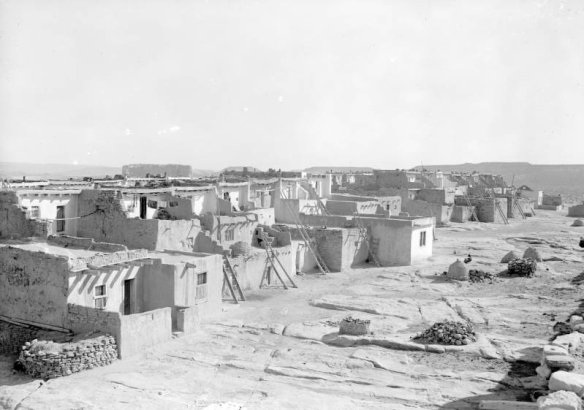 A row of small buildings of the Laguna Pueblo in New Mexico is shown from the back. Wooden ladders lead to the upper levels of the structures. The windows and doors are framed with wood. The surface of the ground is composed of large slabs of rock. Small mounds, probably ovens, stand a few feet behind the buildings. Additional mesas, including the "Enchanted Mesa," are in the distance.