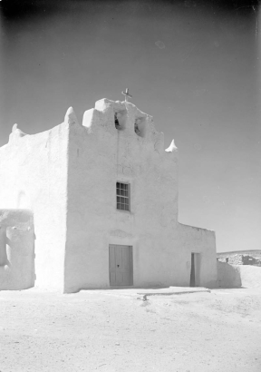 The front of the Church of San Jose at the Laguna Pueblo in New Mexico has a large bell tower with two bells and a large cross. A large window is centered over the large, wooden door; a smaller entrance is in the adjacent one-story section of the church. The adobe surface is cracked in several areas, particularly above the main door and window.  The church is dated at approximately 1699.