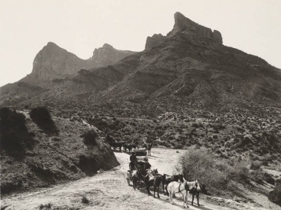 View of wagons, one with a covered wagon frame, pulled by teams of six horses on a dirt road in Arizona. Shows men and supplies in the wagons, rocky buttes, brush and saguaro cacti near the road on the Apache Trail in probably the Superstition Mountains.