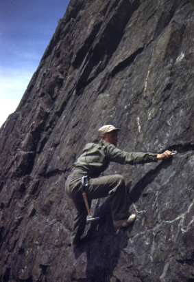Tenth Mountain Division soldier, Joe Stettner, climbs a steep rock face near Camp Hale, Colorado. He wears a cap, khaki uniform, and soft-soled shoes. Hammer and carabiners hang from his belt as he clips a carabiner to a piton.