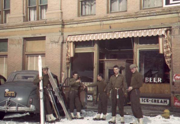 Five Tenth Mountain Division Skitroopers in uniform stand outside a small store at the Jerome Hotel in Aspen, Colorado. Anthony Dillon, far left, holds skis. Man on far right is identified as photographer Bill Southworth.
