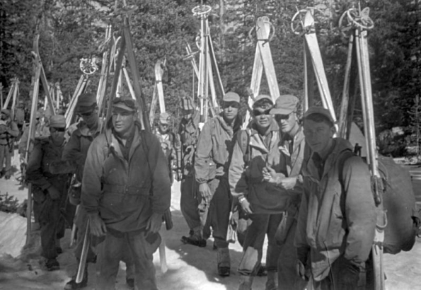 Shows a group of Tenth Mountain Division skitroopers standing on the snow in the mountains. They have their skis and poles attached to the sides of their knapsacks. All of the are wearing caps, anoracks, pants and boots with gaiters. Four of them are also wearing ski goggles.
