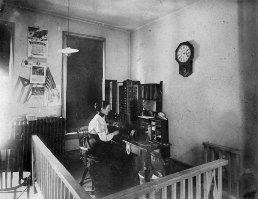 A woman telephone operator is sitting at the first switchboard in  Idaho Springs, Colorado. The headset she is wearing has a transmitter at mouth level, and wires connected to the board. Her surroundings include: an  enclosing wooden railing with a single swinging gate, three calenders on the wall, two flags, a radiator with books on it, and a clock which reads 2:16. Electric lights hang overhead.