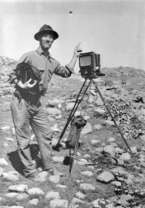 Jesse Nusbaum stands next to a camera tripod and leans his hand on top of it pointing his index finger up. He holds a bag and some film cartridges in his other hand and has a cigarette in his mouth. He wears wire-framed glasses and a hat. A cloth is tied to one leg of the tripod.