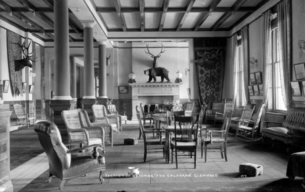 Eastward view of the rotunda (lobby) of the Hotel Colorado in Glenwood Springs, Colorado; various upholstered, wicker, and wooden chairs and tables; three supporting pillars at left; two windows at right; fireplace at eastern wall; taxidermied animals mounted on walls.