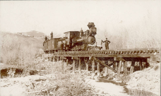 View of a Denver and Rio Grande Railroad train at Maysville (Chaffee County), Colorado.  Shows locomotive number 52 and box cars on the bridge at the North Fork of the South Arkansas River. Men in work clothes stand on and near the train, snow covered mountains are in the distance.