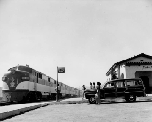 View at the Southern Pacific Railroad depot in Palm Springs (Riverside County), California. Women pose by a 1948 Ford "woody" station wagon and the Mission Style building; the Southern Pacific locomotive and streamliner "Golden State" waits by a sign that reads: "Palm Springs Stage."