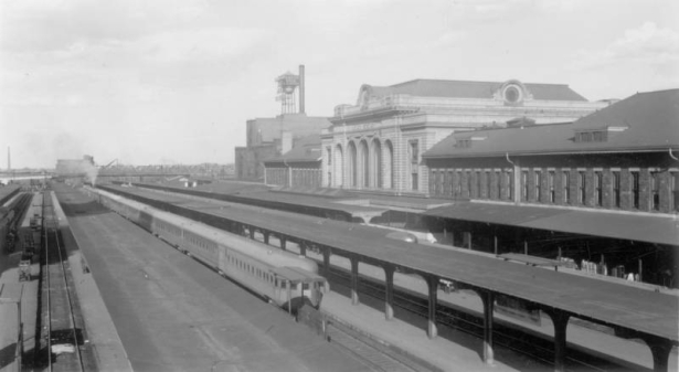 View of the Union Pacific "City of Denver" and the Chicago, Burlington & Quincy Railroad "Denver Zephyr" trains, umbrella sheds and passenger platforms at Union Station in Denver, Colorado. The railroad depot is a rusticated stone building. The 20th (Twentieth) Street viaduct is in the distance.