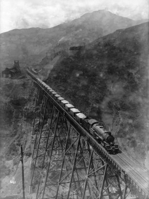 View of a steel Bingham & Garfield Railroad trestle over Markham Gulch in Bingham Canyon, Salt Lake County, Utah. A Bingham & Garfield 0-8-8-0 locomotive number "101" hauls copper ore on the trestle. Shows wooden Utah Copper Company (later Kennecott Copper Corporation) buildings, probably tramway cables and a tower. The Oquirrh Mountains are in the distance.