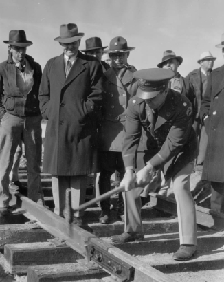 View of a dedication ceremony with men in military uniforms, or suits and overcoats, at Lowry Army Air Force Base in the Lowry Field neighborhood of Denver, Colorado. The men watch an officer hammer a spike into railroad tracks constructed by the Work Projects Administration (WPA)  The men wear caps and fedoras. The tracks were constructed to connect Lowry to the Union Pacific Railroad line.