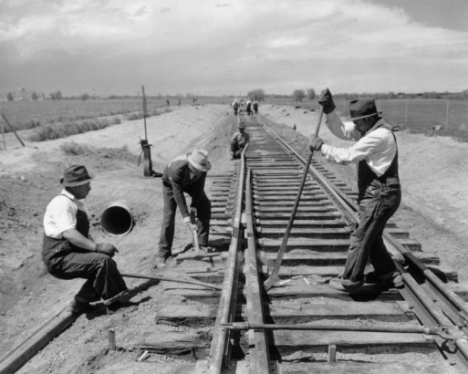 Work Projects Administration (WPA) railroad construction workers in overalls and fedoras use a hammers and a crowbar to install rails at a switch on railroad tracks near Lowry Air Force Base in the Lowry Field neighborhood of Denver, Colorado. Shows a railroad signal and a culvert beside the tracks. The tracks were constructed to connect Lowry to the Union Pacific Railroad line.