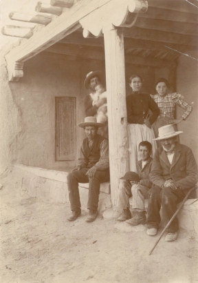 Portrait of a Spanish American family in front of their adobe house in Northern New Mexico. Shows vigas, decorative wooden posts, and shutters. The women wear simple blouses and skirts with their hair pulled back and the men wear vests or coats with hats. One man holds a baby and an elderly man, with a white beard, sits with his cane beside him.