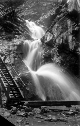 View from the base of Three Falls waterfall in South Cheyenne Canyon, southwest of Colorado Springs, Colorado; wooden walkway crosses pool in foreground; stairs are parallel to waterfall.