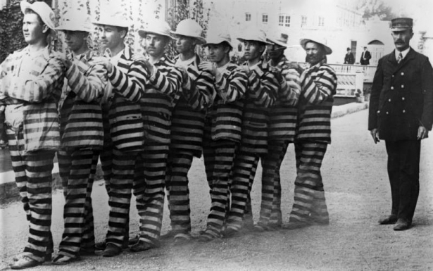 Convicts at the State Penitentiary in Canon City, Colorado, wear uniforms marked with wide, horizontal stripes. They stand in a line, each with his hands on the shoulders of the man in front of him. A uniformed guard in a double breasted jacket and conductor's hat looks on.