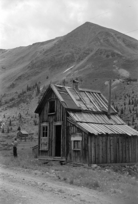 An unidentified woman stands at the side of a large, two-story log cabin in Chihuahua Gulch near Montezuma in Summit County, Colorado. A dirt road runs from the foreground past the cabin. Pine trees stand at the base of a mountain ridge in the background.