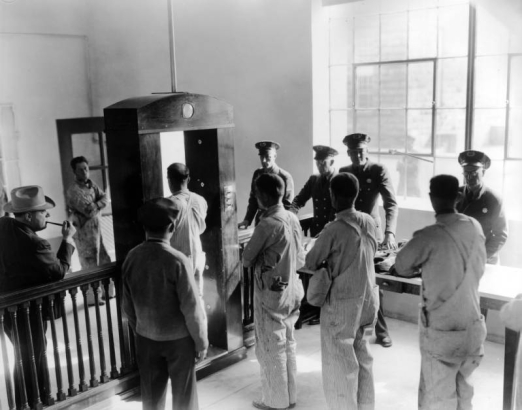 Inmates at the State Penitentiary in Canon City, Colorado, wear pinstriped bib overalls and stand with arms crossed as they wait to pass through a wood electric eye gate flanked by spindle balustrades. Uniformed guards and a pipe smoking man in a suit look on; factory windows provide interior lighting.