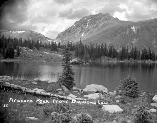 Two pine trees stand among large rocks on the near shore of Diamond Lake in Boulder County in This view looking toward Arapaho Peak in the distance on the border of Boulder and Grand counties in Colorado. Small  patches of snow are on Arapaho Peak.