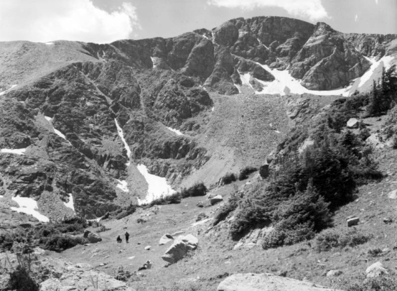 A group of three unidentified people is in the left foreground in the Mammoth Basin west of Apex in Gilpin County, Colorado. One person sits alone and the other two are farther away, one sitting on a rock, the other standing. In the distance, the rocky ridge of James Peak has small patches of snow on it. James Peak stands where Gilpin, Grand and Clear Creek Counties converge in Colorado.