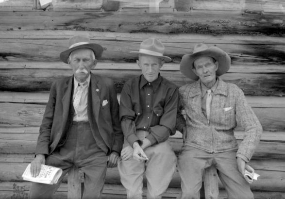 John A. (Jack) Gilfillan, John H. Kemp (father of the photographer) and J. B. Rowley sit on a log railing in front of a log cabin  in Eldora, a mining town in Boulder County, Colorado. The three men each helped found the town in the late 1890s. All three wear hats. Gilfillan and  Rowley hold newspapers in their hands, and Kemp has a cigarette in his right hand.