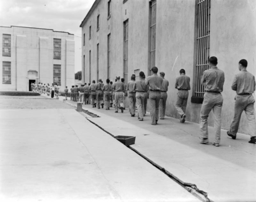 Convicts march with arms crossed at the State Penitentiary in Canon City, Colorado. The concrete wall next to them has barred windows. The inmates approach cell house #1, a three-story concrete building with barred windows.