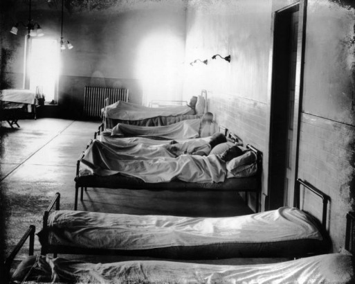 Black and white convicts lie in cots in the hospital at the State Penitentiary in Canon City, Colorado. The walls are partly tiled; electric lights and a steam radiator furnish the interior.