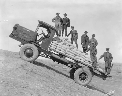 View of a Coleman flatbed truck on an incline with the city of Denver, Colorado, in the distance. A group of well-dressed men and workers ride on the truck loaded with sacks of probably sand. The surrounding landscape is barren. The state capitol building is visible in the distance.