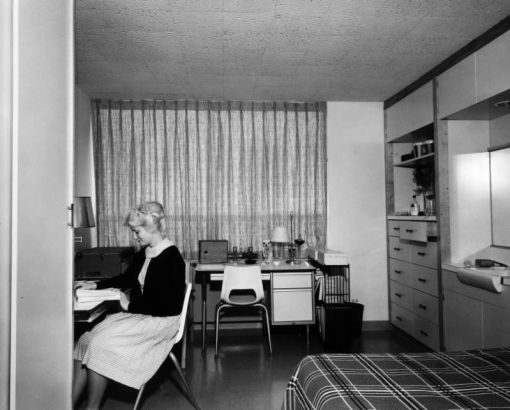 Interior view of a dormitory room in the Escalante Terrace Western State College, Gunnison, Colorado; Berne, Muchow, Baume & Polivnck, architects; a woman sits reading at a desk; a plaid bedspread covers the bed, built-in drawers, shelves and sink are along one wall with bongo drums, vase of flowers, bottle of skin lotion; another desk has a lamp, speaker from a record player and knicknacks.