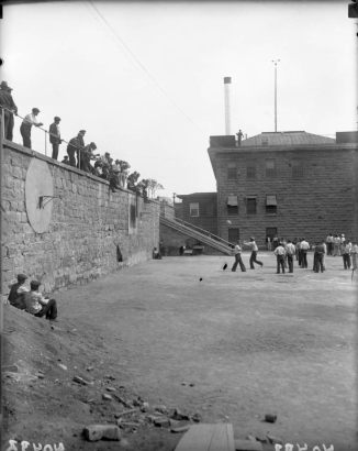 Convicts play basketball at the State Penitentiary in Canon City, Colorado. An armed guard watches from the metal roofed admissions building, with its anemometer. Hoops and backboards are mounted on the rock wall, and prisoners sit on top and lean on the railing. Stairs connect the two levels. Men sit on a dirt pile in the foreground.  Metal smokestack is in the background.