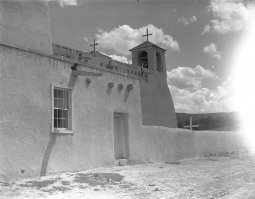 View from the side of the adobe St. Francis of Assisi church in Ranchos de Taos (Taos County), New Mexico. Shows an adobe wall with a doorway and window, wooden vigas, and two bell towers with crosses on top.